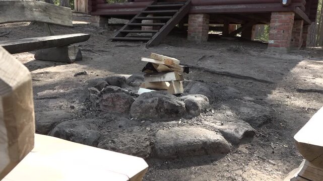 Stacked wooden logs arranged in a circular stone fire pit surrounded by dirt and wooden benches in a forested outdoor setting near a cabin