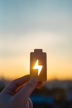 Renewable energy concept showing hand holding wooden battery icon with lightning bolt cutout against sunset sky. Sustainable power, solar charging, and eco-friendly technology solutions.