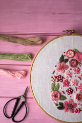 Floral embroidery in wooden hoop with threads, scissors and linen fabric on pink wooden background . Handmade needlework, DIY craft hobby, sewing and textile art concept.