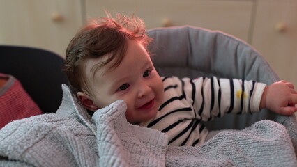 Close-up of baby smiling with hand on soft blanket, joyful and playful expression, sitting comfortably, engaging with surroundings, soft textures and gentle moments
