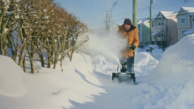 Close-up action: determined man maneuvers wheeled snow blower to remove snow from backyard and walkway. Snow sprays in arcs, realistic winter chore in suburban yard, slow motion details.