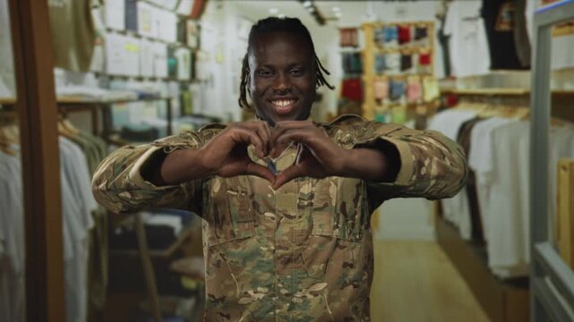 Man soldier wearing camouflage uniform and dog tags forms a heart with hands while smiling in a retail clothing store building lined with racks; pride duty.