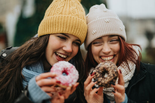 Two teen girls laugh and share donuts, capturing a cheerful, friendly moment in winter outdoors.