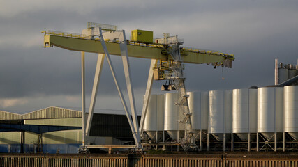 large yellow gantry crane at an industrial port facility next to white silos © Flowal93