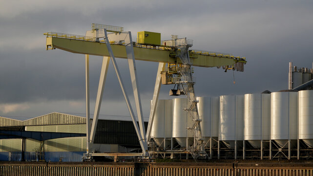 large yellow gantry crane at an industrial port facility next to white silos