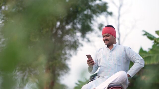 young indian farmer using smartphone sitting at green agricultural field