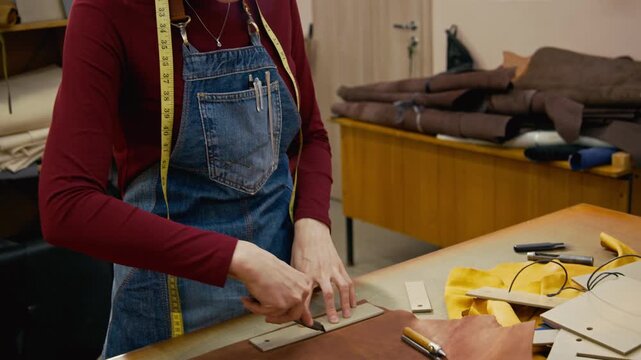 Tilt down shot of young craftswoman cutting brown leather with craft knife tracing acrylic template on workbench while creating handmade wallet in workshop