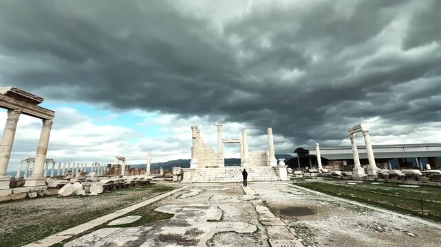 A woman in pants walks among ancient ruins and stops at a column gate at Laodikeia under a cloudy sky.