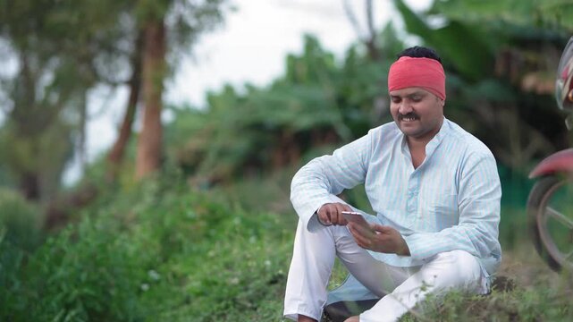 young indian farmer using smartphone sitting at green agricultural field