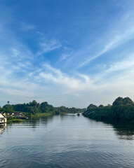 Obraz premium Calm river flowing between lush green banks with riverside houses under a wide blue sky with soft clouds.