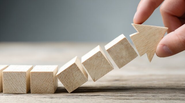 Wooden blocks arranged in an upward staircase leading to an arrow are manipulated by a human hand