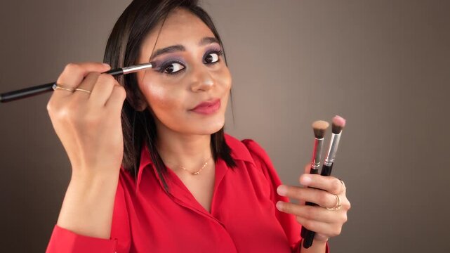 Closeup view of beautiful young woman doing applying eyeshadow make-up looking at camera holding eye makeup brushes in hand, isolated over grey studio background. Cosmetic brushes advertising concept.