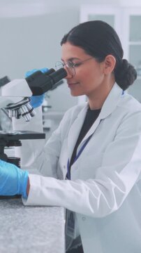 Woman scientist of Hispanic descent calibrates microscope to analyze slide within sterile laboratory setting