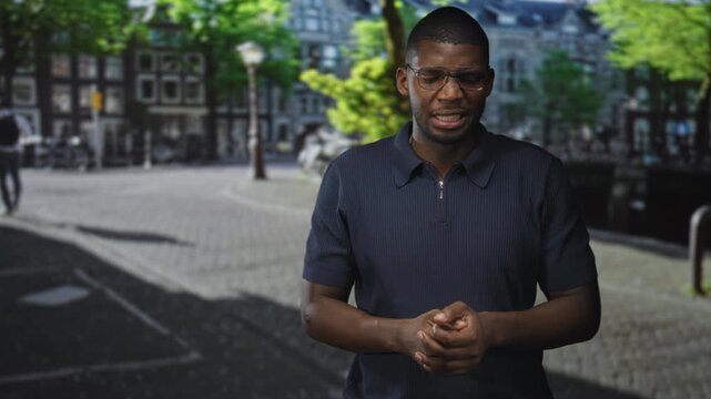 Young black man rubs and clasps hands while speaking on a cobbled canal side city street with lamp post; contemplative.