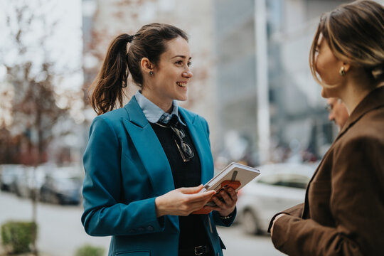 A smiling woman in a blue blazer chats with a colleague outdoors, holding a notebook. Two professionals converse on a city street, capturing a casual business moment.