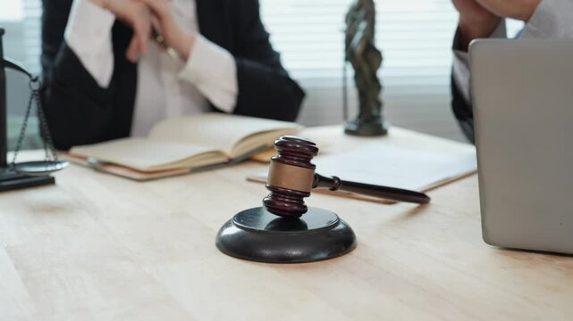 An Asian businessman and a female lawyer, both in suits, are examining documents with hammers at a wooden table near a window in an office, indicating that legal proceedings are underway.