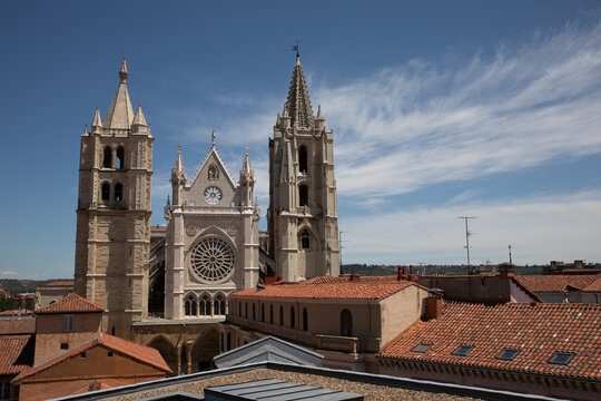 View of the Gothic cathedral's towers and rose window rise above the red-tiled rooftops under a bright blue sky streaked with clouds, Leon, Castile and Leon, Spain.