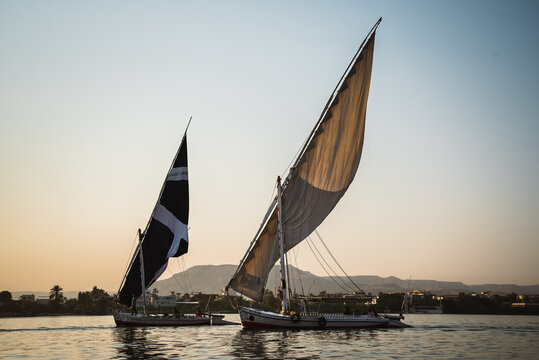 View of feluccas with billowing sails glide across the tranquil waters, their reflections shimmering in the fading light, Luxor, Luxor Governorate, Egypt.