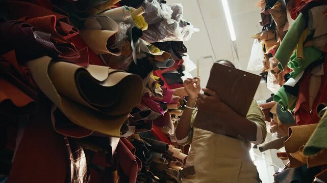 Low angle shot of young African American female artisan in apron writing notes on clipboard while counting hides and sides organizing leather stock in workshop storage room