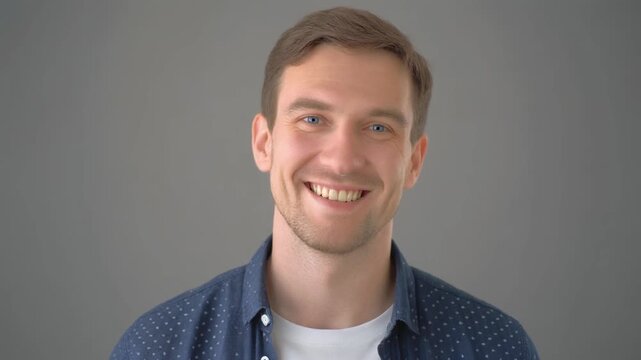 Portrait of a smiling young Caucasian man with blue eyes wearing a blue patterned shirt over a white t-shirt. Friendly facial expression on a neutral grey background.