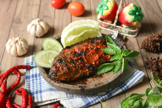 A plate of Indonesian-style spicy grilled tilapia, known as ikan bakar bumbu rujak, served with fiery sambal and fresh raw vegetables like cabbage, cucumber, and lemon basil.