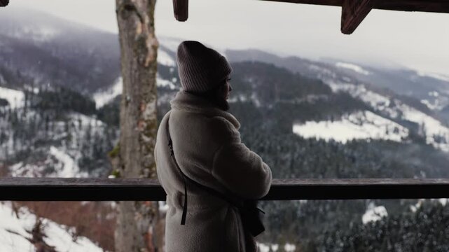 A woman steps into the frame onto a wooden balcony and pauses to admire the wide snowy mountain landscape. She stands quietly, looking across the forested hills and winter valleys, enjoying the peace