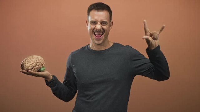 Hispanic man holding brain model with tongue out and right hand showing rock horns in studio; playful curiosity.