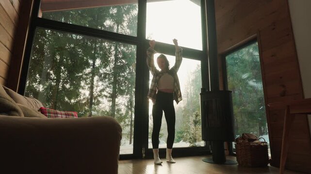 A young woman dances freely in a warm wooden cabin filled with soft natural light. She moves playfully near a large panoramic window, with tall trees and a snowy forest visible outside.
