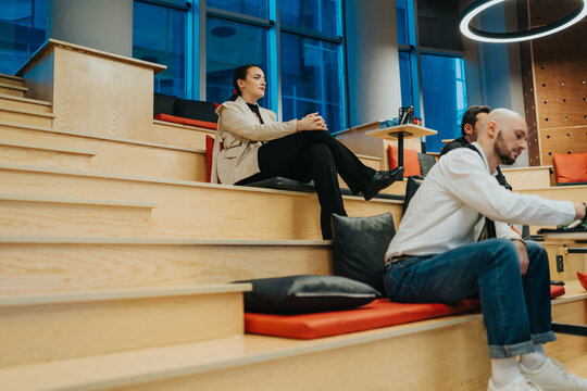 A woman sits on wooden steps in a bright coworking space while colleagues talk nearby. Casual scene of professionals relaxing on tiered seating with cushions and contemporary interior.