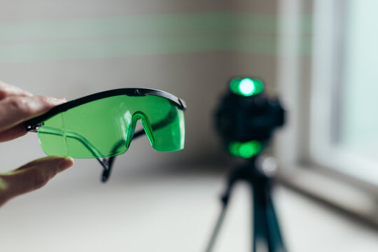 Hand Holding Green Safety Glasses With Laser Level Visible Behind. Personal Protective Equipment In Work Context.
