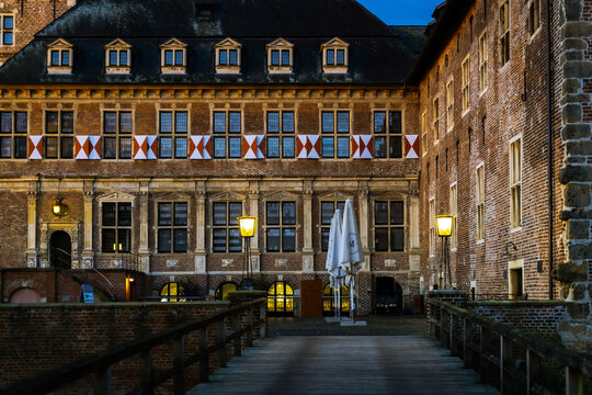 Eine einladende Holzbr&uuml;cke f&uuml;hrt direkt auf die festlich erleuchtete Backsteinfassade des Schlosses zu, deren Fensterl&auml;den in leuchtendem Rot und Wei&szlig; einen starken Kontrast zum tiefblauen Abendhimmel
