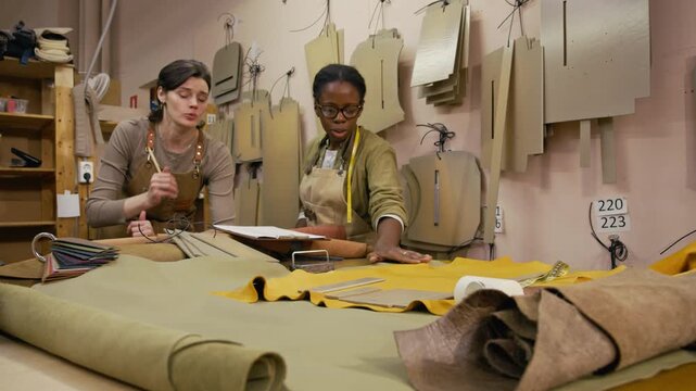 Focused young African American craftswoman discussing leather item design on clipboard with female colleague while choosing material on workbench in leathercraft studio