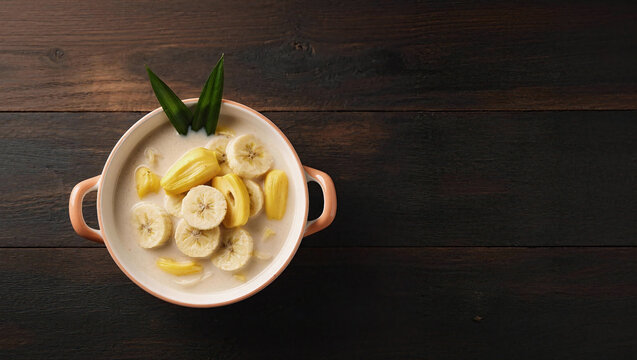 A traditional Southeast Asian sweet dessert featuring sliced ​​bananas and chunks of yellow jackfruit called Kolak served in thick coconut milk, garnished with pandan leaves in a ceramic bowl with a h