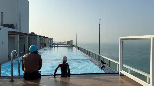 Rooftop infinity pool man and child sit at edge overlooking calm ocean at sunrise, adult guiding young swimmer with caps, quiet