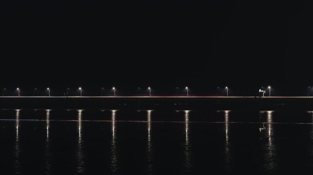 A long bridge with repeating streetlights at night reflected in the water. Atmospheric, dark