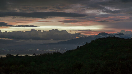 Fototapeta premium Layered clouds stretch across the evening sky above dark forested hills, with distant coastal water and scattered lights visible below. 