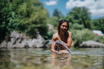 Fototapeta premium A woman smiles and splashes water while standing in a shallow riverside. Bright outdoor scene with trees, rocks, and a blue sky.