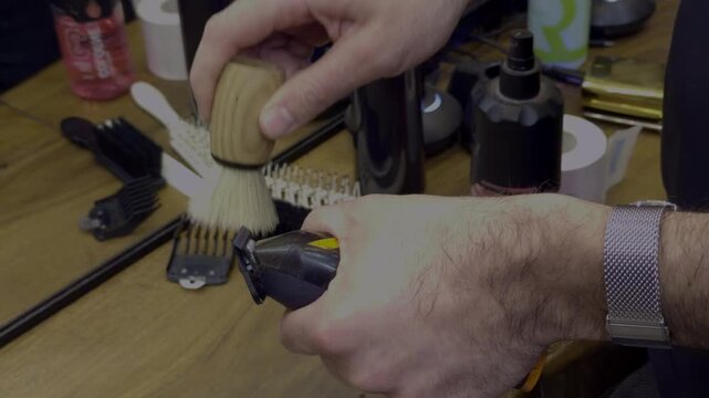 Close-up of a professional barber meticulously maintaining equipment by using a soft-bristle wooden brush to sweep hair debris from electric trimmers and applying disinfectant spray over a workstation