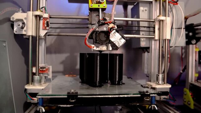 A 3D printer in operation, printing plastic parts using fused filament manufacturing technology. The print head deposits molten material onto a glass work plate, forming detailed black components.
