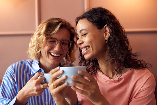 Two diverse women enjoying coffee, from a blue coffee mug, and conversation in a cozy cafe, sharing smiles