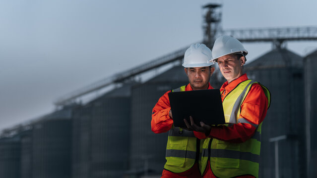 Two male industrial engineers wearing helmets are discussing collaborating on a new project. The engineering team is monitoring a gas separation plant using a laptop.