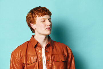 Obraz premium Young man with red hair and freckles wearing brown leather jacket stands with closed eyes against blue background for portrait and fashion concept.