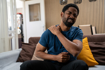 Fototapeta premium Black man looking uncomfortable with shoulder pain in living room
