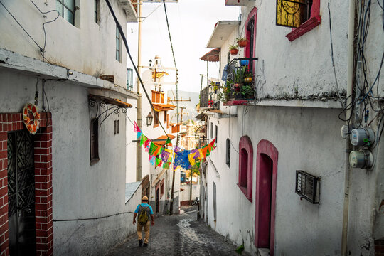 Local man with backpack walking down narrow cobblestone street with colorful papel picado banners toward historic center in Taxco, Mexico
