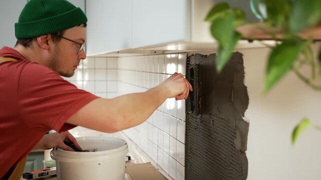 Professional tiler meticulously applying dark gray tile adhesive with a notched trowel to a kitchen wall, preparing the surface for white ceramic tile backsplash installation