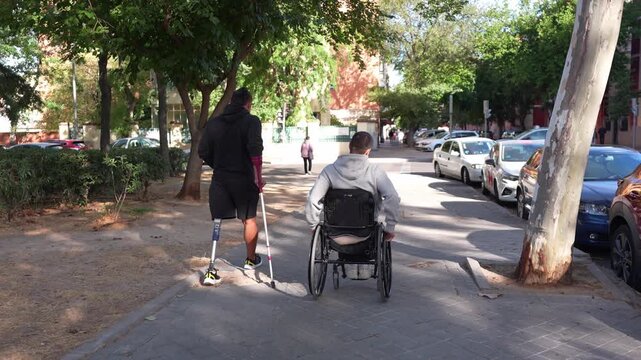 Two disabled friends sharing an inclusive moment. An amputee man with a prosthetic leg and another in a wheelchair walking on a city sidewalk