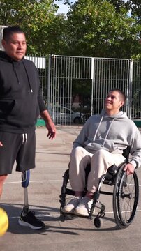 Two male friends with disabilities enjoying a game of basketball outdoors. One man has a prosthetic leg, and the other is in a wheelchair