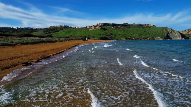 Aerial drone view of the most beautiful Malta beach in Gozo, vibrant red sand Ramla beach with crystal clear blue water and mesmerizing waves with lush green vegetation around it and the famous Mixta 