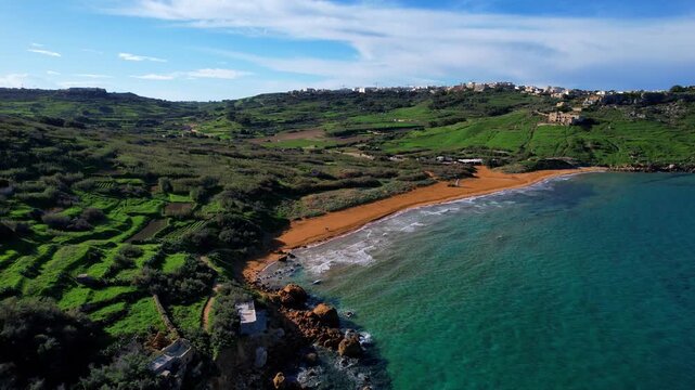 Aerial drone view of the most beautiful Malta beach in Gozo, vibrant red sand Ramla beach with crystal clear blue water and mesmerizing waves with lush green vegetation around it and the famous Mixta 