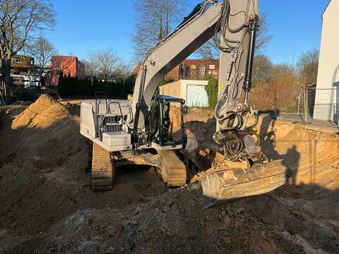 Large tracked excavator operating on a residential construction site, digging deep foundation trenches in sandy soil under clear blue sky.